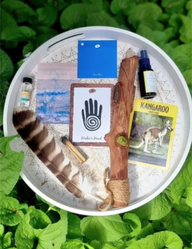 Ceremonial tools used at Hill Tribe Healing, including a feather, oracle cards, a wooden smudging tool and an essential oil spray, arranged on a decorative tray surrounded by green leaves.