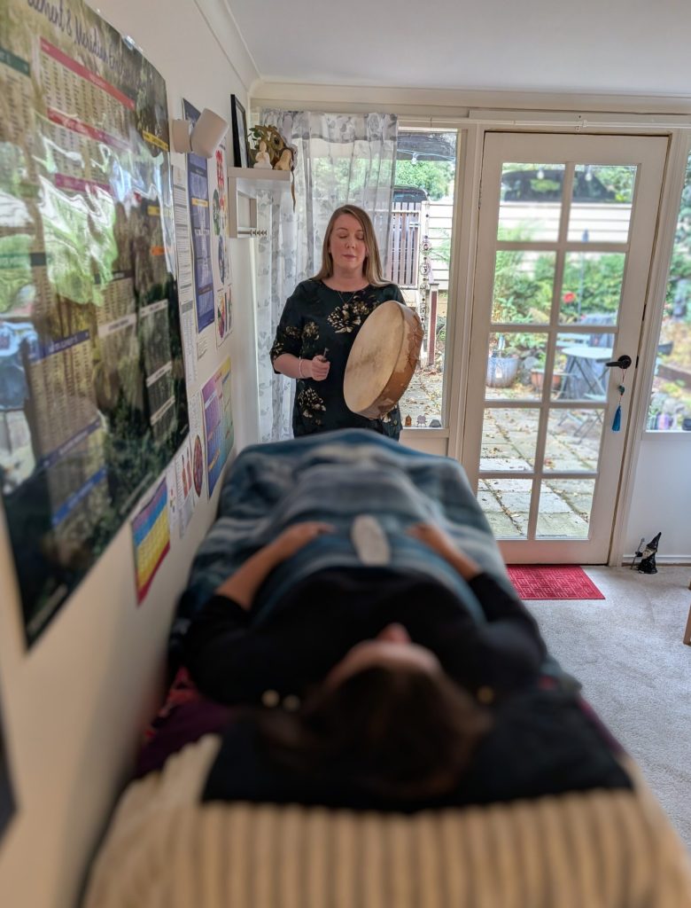 Kath Whitehorn leading a one on one session while holding a frame drum, with a client resting comfortably on a treatment table at Hill Tribe Healing.