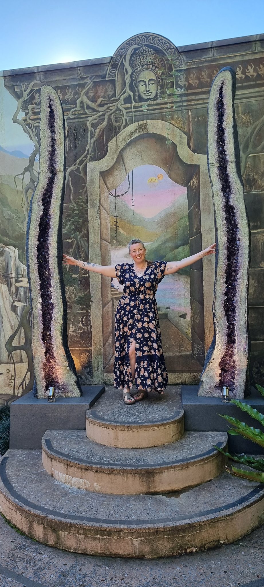 Kath Whitehorn standing between two large amethyst crystal formations in front of a decorative stone archway, smiling with her arms outstretched.